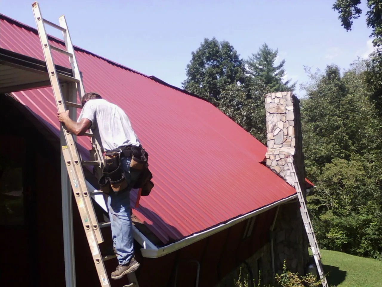 Roofer on a ladder beside a red metal roof and stone chimney — Soffit & Fascia Repair by Roofing Brothers in Green Isle
